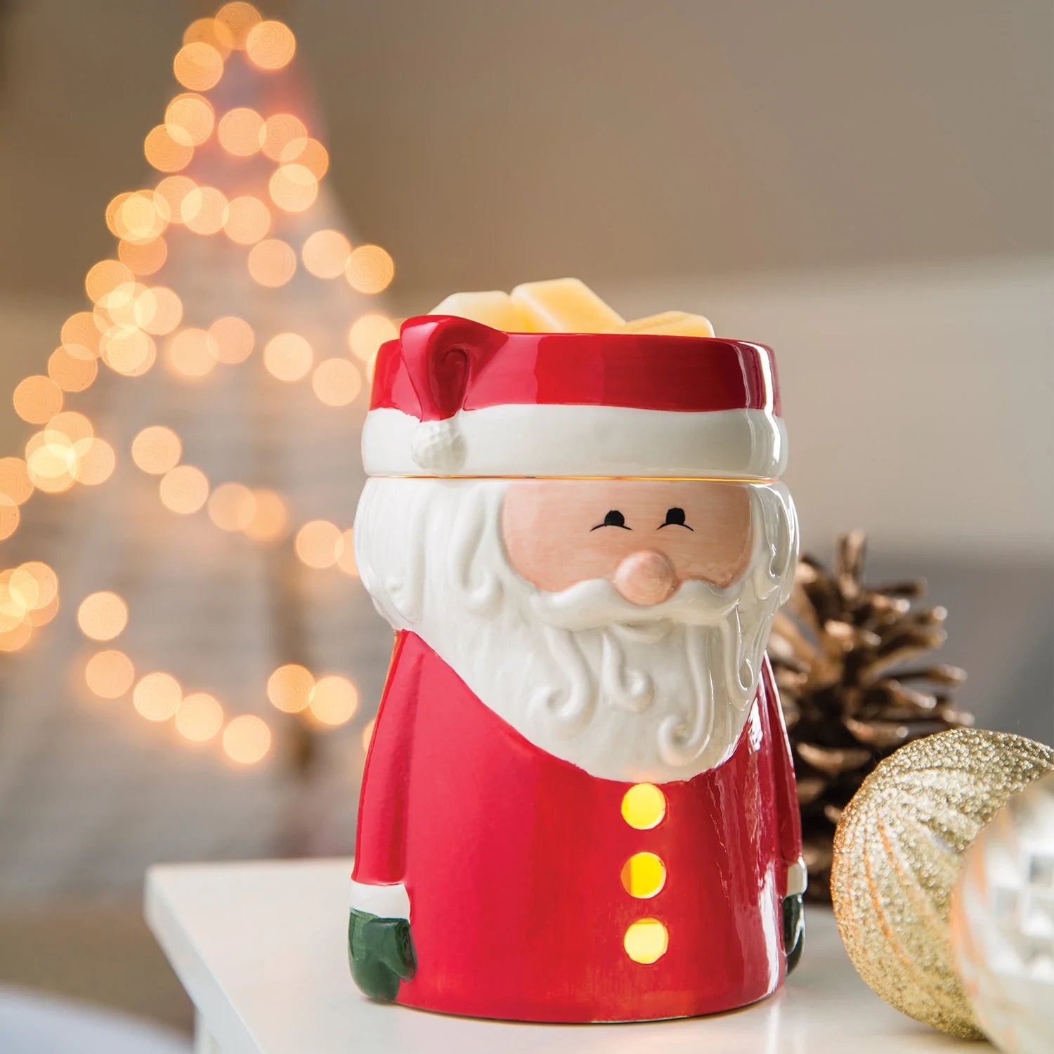 Ceramic Santa-themed electric wax melter on a table surrounded by baubles, with blurred Christmas tree lights in the background