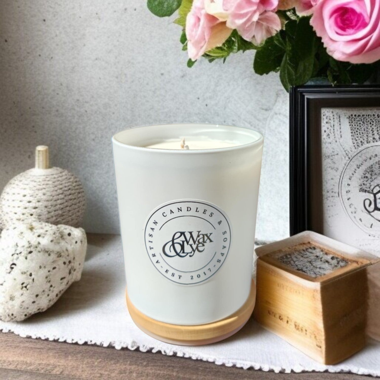 White glass candle with Wax & Lye logo resting on its timber lid, placed in front of a stylised background.